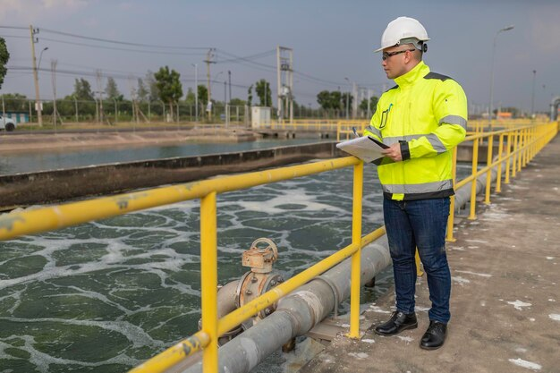 An engineer wearing a high-visibility jacket, safety helmet, and holding a clipboard stands by a wastewater treatment facility, observing the water processing area. The scene highlights industrial water treatment and environmental monitoring.