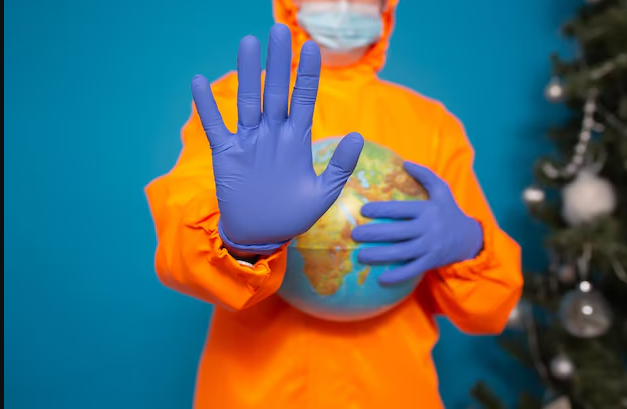 A person in an orange hazmat suit and face mask holding a globe with one hand while raising the other hand wearing a blue glove in a "stop" gesture. The background is blue with a partially visible Christmas tree on the side.