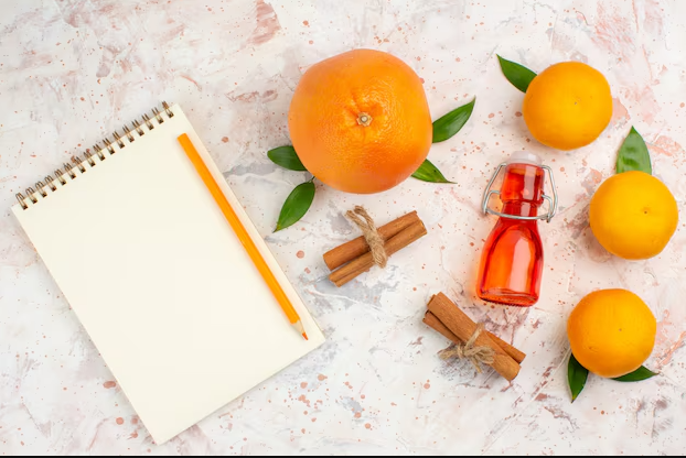 A flat lay composition featuring a blank notebook with an orange pencil on the left, surrounded by fresh oranges, cinnamon sticks tied with twine, green leaves, and a small glass bottle filled with orange-colored liquid on a textured background.
