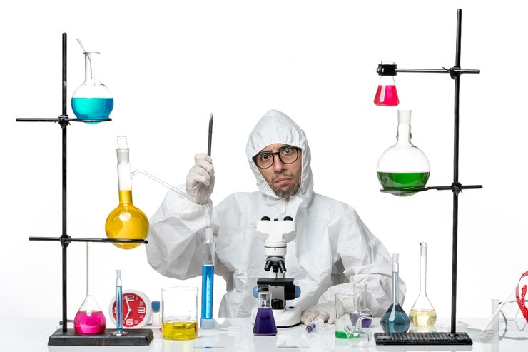 A scientist wearing a white lab suit and gloves is standing at a laboratory bench with various colorful chemical solutions in flasks. The scientist is holding a tweezers in one hand, and there is a microscope in front of him. There are also several glass containers and colorful liquids around the workspace.