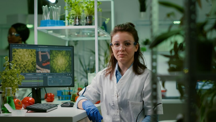 A female scientist in a lab coat and safety glasses is sitting at a laboratory desk. She is wearing blue gloves and looking directly at the camera. Behind her, there are plants and a computer screen displaying images of microscopic plant cells.